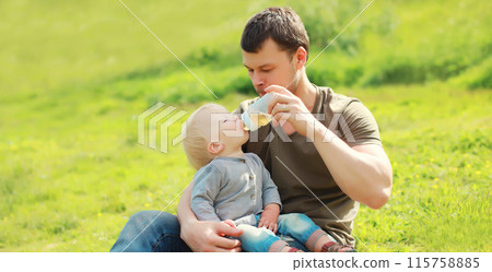 Father feeds baby from bottle sitting on grass in sunny summer park 115758885