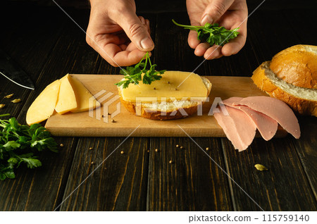 Making sandwiches with cheese and parsley. Close-up hands of a chef on the kitchen table preparing breakfast. Place for advertising on a dark background Making sandwiches with cheese and parsley. Close-up hands of a chef on the kitchen table preparing breakfast. Place for advertising on a dark background 115759140