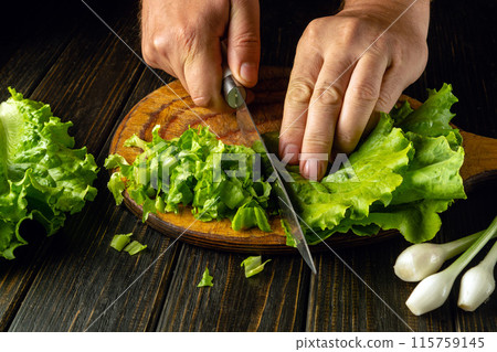 Chef cutting lettuce leaves on wooden board with knife in hand before preparing tasty breakfast. Vegetable diet concept 115759145