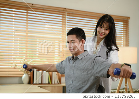 Shot of smiling female rehabilitation specialist assisting man with dumbbell exercises at clinic 115759263