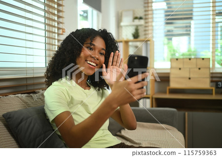 Cheerful young African American woman waving hand making video call on smartphone at home 115759363
