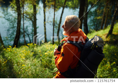High angle view of senior male tourist wearing orange jacket with gray backpack looking at forest lake, copy space 115761754