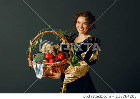 Portrait of a young adult woman dressed in a medieval dress holding a basket with vegetables and fruits, apples and brussels sprouts. Harvest concept. 115764205
