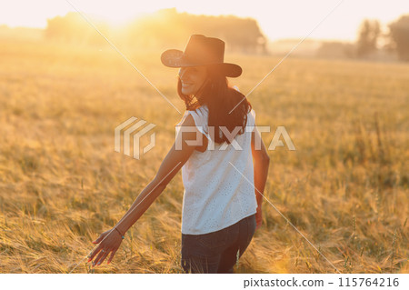 Woman farmer in cowboy hat walking with hands on ears at agricultural wheat field on sunset. Woman farmer in cowboy hat walking with hands on ears at agricultural wheat field on sunset. 115764216