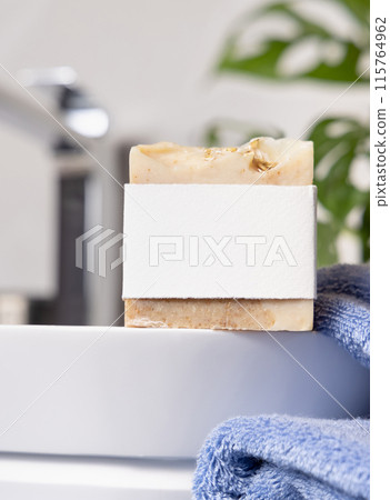 Soap bar with blank label on vessel sink near blue towels and plant close up, mockup 115764962