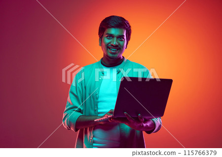 Young, attractive Indian man looks at camera while using laptop in neon light against gradient studio background. 115766379