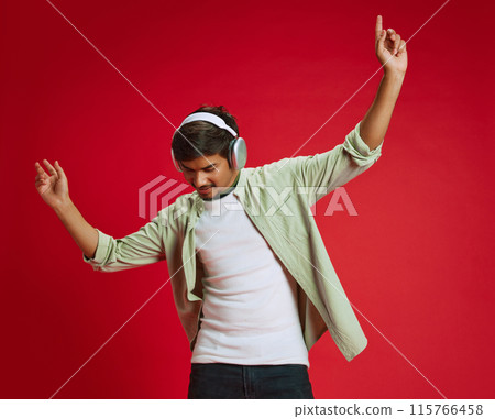 Portrait of young Indian man dressed casual attire and dancing while listening music in headphones against red studio background. 115766458