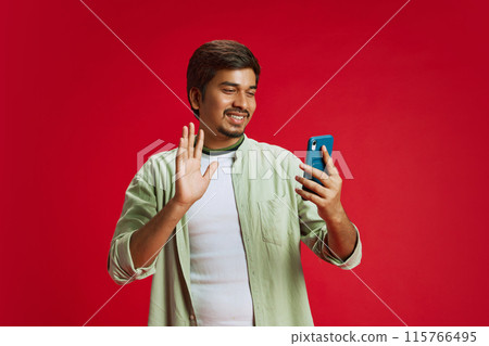Attractive Indian man gesturing hello while talking, using phone for communicate in distance against red studio background. 115766495