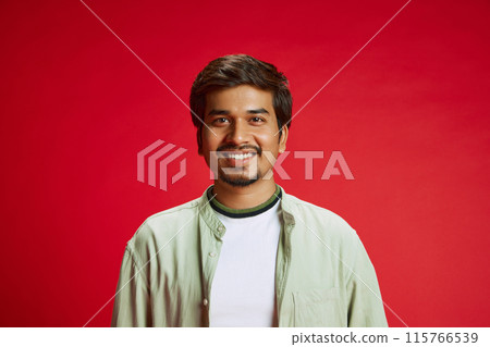 Smiling young Indian man, student in casual attire looking at camera against red studio background. 115766539