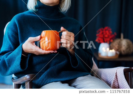 Cropped mature middle aged woman in sweater and warm socks relaxing in armchair with pumpkin shaped cup of hot coffee or tea drink. Cozy calm autumn holidays at home. Fall hygge mood concept 115767858