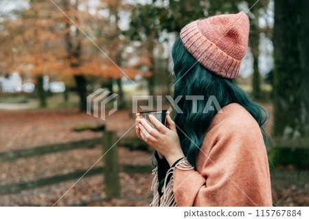 Side view Woman in hat wrapped in Shawl Poncho drinking hot drink from thermos cup during autumn walk in forest with fall leaves. Feeling harmony, relax, personal fulfillment. Local travel lifestyle. 115767884