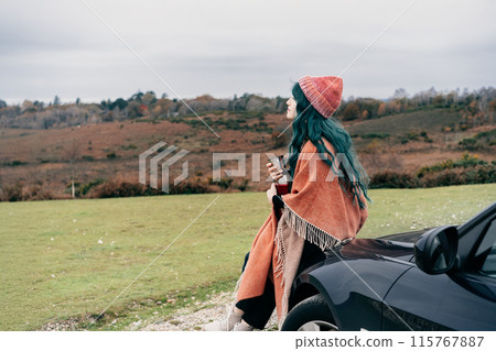 Hipster Woman sits on car's hood, wrapped in warm Shawl Wraps Poncho Cape and hat and enjoying warm drink, while overlooking rural landscape during road trip. Cold season travel, vacation concept Hipster Woman sits on car's hood, wrapped in warm Shawl Wraps Poncho Cape and hat and enjoying warm drink, while overlooking rural landscape during road trip. Cold season travel, vacation concept 115767887