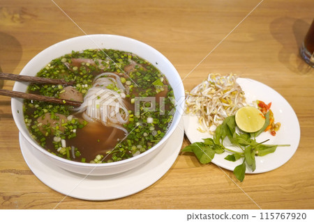 A bowl of pho on a wooden table. The pho appears to have rice noodles, sliced beef, and a clear broth. A bowl of pho on a wooden table. The pho appears to have rice noodles, sliced beef, and a clear broth. 115767920