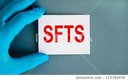Hand in blue glove with white card. Concept word 'SFTS'. COVID-19 Pandemic Coronavirus concept. Beautiful blue background. Hand in blue glove with white card. Concept word 'SFTS'. COVID-19 Pandemic Coronavirus concept. Beautiful blue background. 115768456