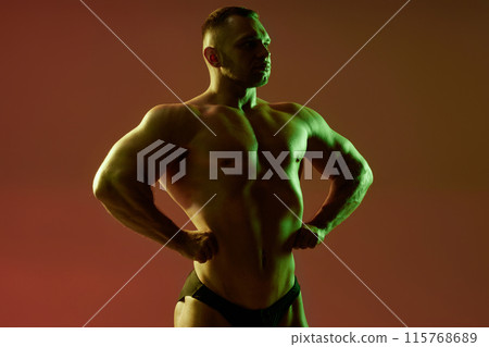 Studio image of young male bodybuilder in front lat spread pose showcasing chest and abs muscles relief. Medium shot of athlete in contour green and neutral light on red background, copy space Studio image of young male bodybuilder in front lat spread pose showcasing chest and abs muscles relief. Medium shot of athlete in contour green and neutral light on red background, copy space 115768689