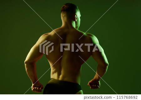 Studio medium shot of pumped up back of unrecognizable brutal male bodybuilder spreading wide shoulders showing flexed muscles in contour green and neutral light, copy space Studio medium shot of pumped up back of unrecognizable brutal male bodybuilder spreading wide shoulders showing flexed muscles in contour green and neutral light, copy space 115768692