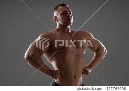 Medium backlight shot of topless strong male bodybuilder with hard chest muscles in front lat spread pose showing wide shoulders in shady studio with dramatic light on grey background, copy space 115768698