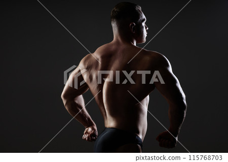 Medium backlight image of sexy brutal male model sideways to camera showing flexed back muscles in dark studio with dramatic light on black background, copy space Medium backlight image of sexy brutal male model sideways to camera showing flexed back muscles in dark studio with dramatic light on black background, copy space 115768703