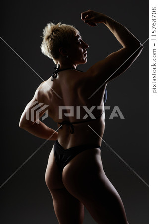 Vertical backlight shot of athletic woman in rear lat and biceps pose showcasing musculature. Medium full profile shot of female bodybuilder in dark studio with dramatic light on black background Vertical backlight shot of athletic woman in rear lat and biceps pose showcasing musculature. Medium full profile shot of female bodybuilder in dark studio with dramatic light on black background 115768708