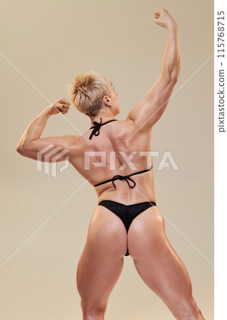 Vertical rear view shot of energetic female bodybuilder striking contest pose showing back and arm muscles isolated in studio on neutral background Vertical rear view shot of energetic female bodybuilder striking contest pose showing back and arm muscles isolated in studio on neutral background 115768715