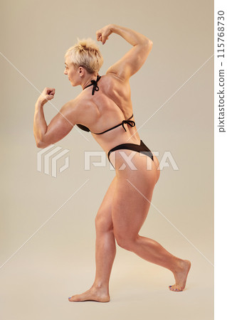 Vertical full shot of bodybuilding female competitor showcasing three quarter back biceps pose while extending broad shoulders contracting biceps isolated in studio on neutral background, copy space Vertical full shot of bodybuilding female competitor showcasing three quarter back biceps pose while extending broad shoulders contracting biceps isolated in studio on neutral background, copy space 115768730