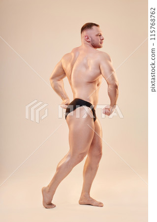 Full body side view shot of handsome bodybuilder topless performing quarter turn pose in studio. Side view of bulked up sportsman demonstrating body musculature on neutral background, copy space Full body side view shot of handsome bodybuilder topless performing quarter turn pose in studio. Side view of bulked up sportsman demonstrating body musculature on neutral background, copy space 115768762
