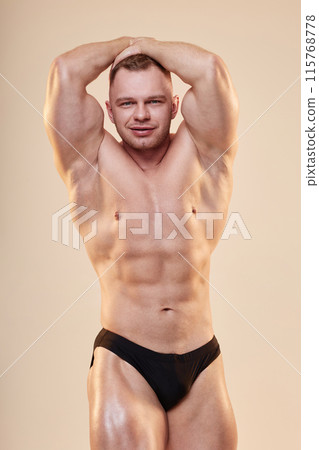Handsome male bodybuilder posing in ab and thigh pose shirtless in briefs showing six-pack isolated on beige neutral background, copy space Handsome male bodybuilder posing in ab and thigh pose shirtless in briefs showing six-pack isolated on beige neutral background, copy space 115768778