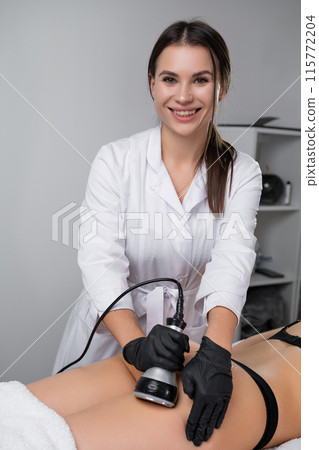 Smiling Cosmetologist Performing Ultrasound Cavitation Treatment on Clients Buttocks in a Modern Clinic 115772204