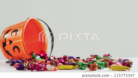 Happy halloween text banner against halloween candies fallen from pumpkin shaped bucket 115773347