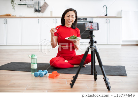 Woman in activewear eating salad while recording fitness video in kitchen Woman in activewear eating salad while recording fitness video in kitchen 115774649