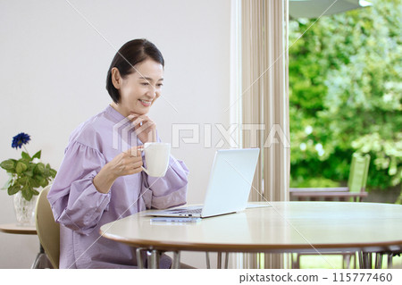 A middle-aged woman drinking coffee and looking at a computer in the living room A middle-aged woman drinking coffee and looking at a computer in the living room 115777460