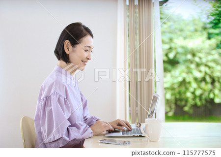 A middle-aged woman drinking coffee and looking at a computer in the living room 115777575