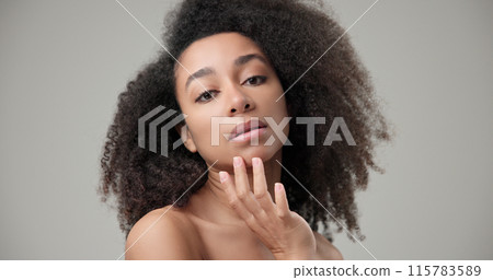 Beauty and healthcare concept - beautiful African American woman with curly afro hairstyle and clean, healthy skin touches her cheek and face with her hand, posing and looking at the camera 115783589