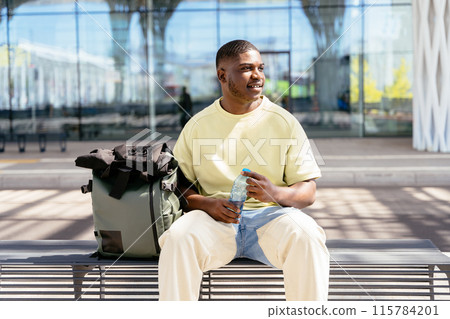 A man sitting on a bench with a backpack and a bottle of water. He is smiling and he is enjoying his time 115784201