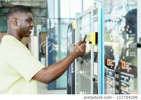 Young traveler black man choosing a snack or drink at vending machine, paying pay pass with smartphone in airport terminal. Vending machine with man. Paying with mobile phone concept. Young traveler black man choosing a snack or drink at vending machine, paying pay pass with smartphone in airport terminal. Vending machine with man. Paying with mobile phone concept. 115784206