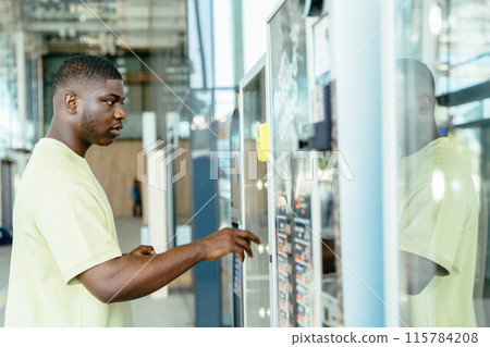 African man in yellow shirt is using a vending machine. The vending machine is located in a public area, and the man is in a hurry. 115784208