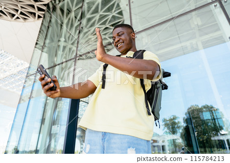 Bottom view traveler African man with backpack taking a picture of himself with his cell phone or having videocall outside at international airport terminal. Air flight or bus trip concept. 115784213