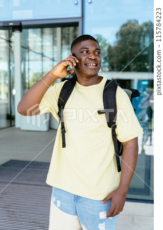 Vertical shot of Nigerian traveler having conversation. Phone call, communication and black man with cellphone in city while on an adventure. Guy with backpack talking on phone on railway station. Vertical shot of Nigerian traveler having conversation. Phone call, communication and black man with cellphone in city while on an adventure. Guy with backpack talking on phone on railway station. 115784223