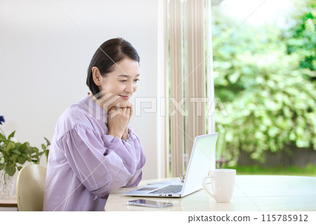 A middle-aged woman drinking coffee and looking at a computer in the living room 115785912