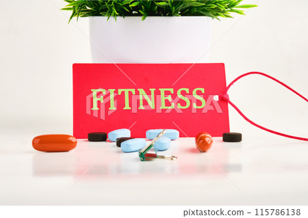Fitness, healthy lifestyle concept. A word FITNESS written on a red card on a white background with pills, vitamins in the foreground Fitness, healthy lifestyle concept. A word FITNESS written on a red card on a white background with pills, vitamins in the foreground 115786138