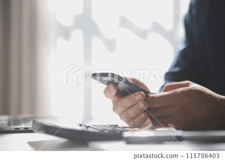 Side view shot of a man's hands using smart phone in interior, rear view of business man hands busy using cell phone at office desk, young male student typing on phone sitting at wooden table, flare Side view shot of a man's hands using smart phone in interior, rear view of business man hands busy using cell phone at office desk, young male student typing on phone sitting at wooden table, flare 115786403