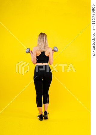 Sporty woman does the exercises with dumbbells on dark, yellow, red background. Sporty woman does the exercises with dumbbells on dark, yellow, red background. 115789909