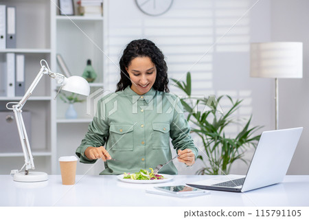 Lunch break at work. Young Latina woman sitting at desk on laptop in office, drinking coffee and eating salad. 115791105
