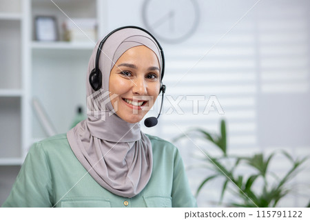Close-up portrait of young Muslim woman wearing hijab and headset standing in office, looking and smiling at camera. Close-up portrait of young Muslim woman wearing hijab and headset standing in office, looking and smiling at camera. 115791122