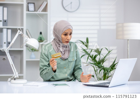 Worried young muslim woman in hijab sitting in office in front of laptop, holding credit card in hand and looking at screen in frustration. Problems with financial account. 115791123
