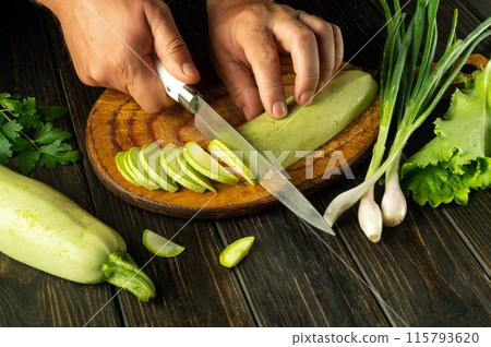 Slicing zucchini with a knife in the hand of a chef for preparing a vegetarian dish on the kitchen table. Advertising space on the black table Slicing zucchini with a knife in the hand of a chef for preparing a vegetarian dish on the kitchen table. Advertising space on the black table 115793620