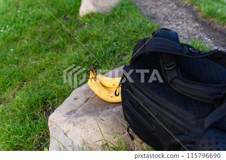 Four ripe bananas lie on a stone next to a black backpack outdoors. 115796690