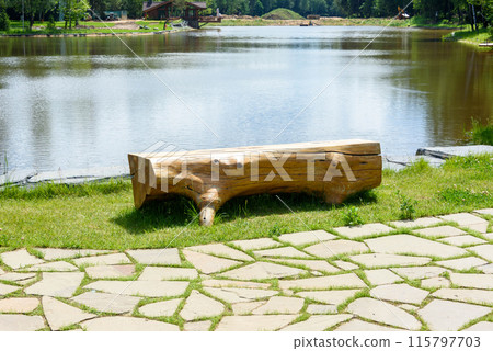 A bench made of a solid tree trunk on the bank of a pond. A bench made of a solid tree trunk on the bank of a pond. 115797703