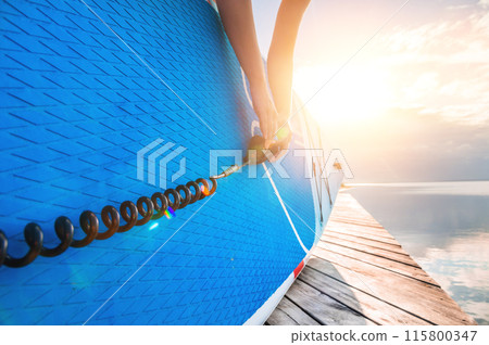 girl stands with an inflatable SUP board and a leash in her hand. Sunset sky in the background 115800347