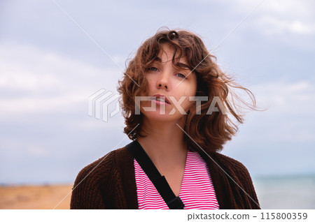 Portrait of a pensive young Caucasian woman with a short haircut. The wind caresses her hair against the backdrop of the beach and sea in cloudy rainy weather Portrait of a pensive young Caucasian woman with a short haircut. The wind caresses her hair against the backdrop of the beach and sea in cloudy rainy weather 115800359